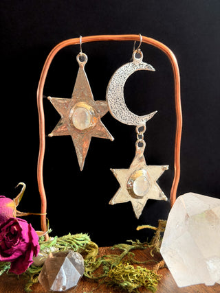 Mismatched star and moon earrings against a black background with flowers, moss and crystals surrounding crafted by Starwitch Metals.