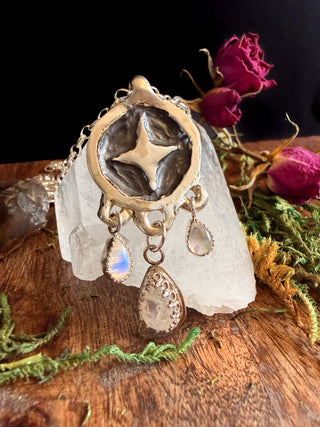 Decorative star pendant with moonstone teardrop dangles on a crystal with dried flowers in the background from Starwitch Metals.